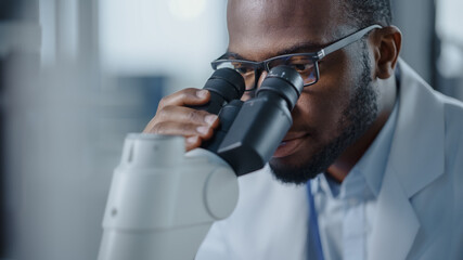 Macro Close Up Shot of a Handsome Black Male Scientist Wearing Glasses and Looking into the Microscope. Microbiologist Working on Molecule Samples in Modern Laboratory with Technological Equipment.