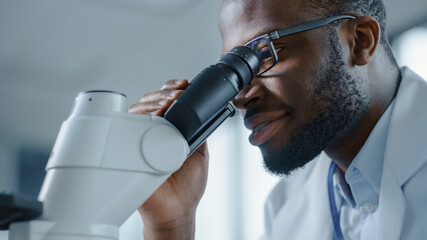Macro Close Up Shot of a Handsome Black Male Scientist Wearing Glasses and Looking into the Microscope. Microbiologist Working on Molecule Samples in Modern Laboratory with Technological Equipment.