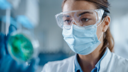 Beautiful Female Scientist Wearing Face Mask and Glasses Looking a Petri Dish with Genetically Modified Sample Chemicals. Microbiologist Working in Modern Laboratory with Technological Equipment.