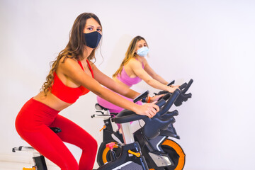 Fototapeta na wymiar Coronavirus pandemic in gyms. Two girls training and having fun on bikes with face masks, social distance and a new normal