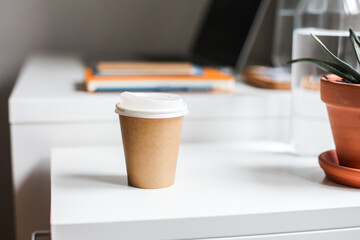 A take away paper cup on a white table. Workspace: laptop, notebooks, a jar of water, a plant