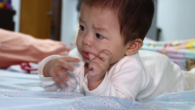 Portrait of 8 month Asian Chinese baby boy lying on his stomach in bed sucking fingers.