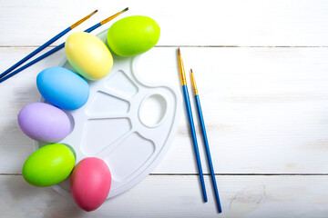 Conceptual photo for the Easter holiday. Painted chicken eggs in a paint palette with paint brushes on a white wooden background with copy space.