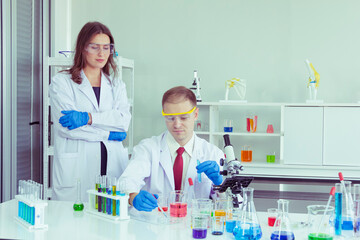 male scientist pours chemicals into a test tube to study chemicals, with a woman scientist serving...