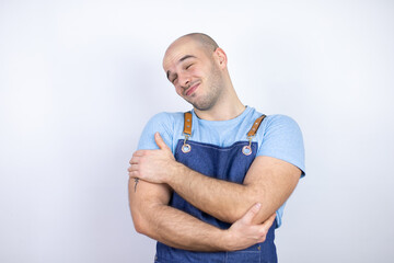 Young bald man wearing apron uniform over isolated white background hugging oneself happy and positive, smiling confident