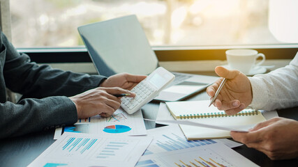 Two businessmen are analyzing cost graphs on their desks in a conference room with a calculator and a laptop.