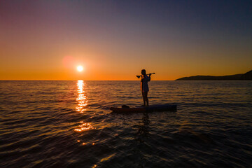 Young woman in two-piece swimsuit and shirt stands on SUP board with oar on her shoulders in the...