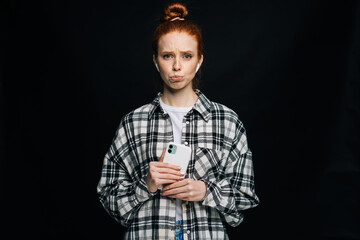 Portrait of sad unhappy young woman holding mobile phone and looking at camera on isolated black background. Pretty redhead lady model emotionally showing facial expressions in studio, copy space.