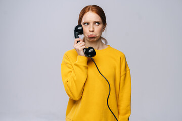 Confused young woman in stylish yellow sweater talking on retro phone and looking away against white background. Pretty redhead lady model emotionally showing facial expressions in studio, copy space