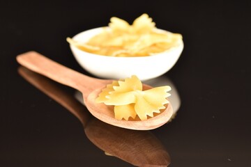 Several yellow pasta, in the shape of a butterfly with a wooden spoon and a white saucer, close-up, isolated on black.