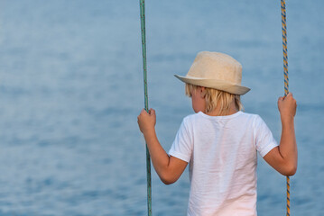 Boy in straw hat riding on a swing overlooking the sea. Happy childhood.