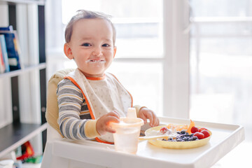 Cute Caucasian baby boy eating ripe berries and fruits with yogurt. Funny smiling child kid sitting...