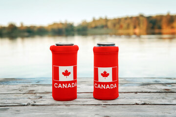 Two cans of beer in red cozy beer can cooler with Canadian flag standing on wooden pier by lake...