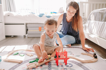 Mother and toddler boy playing with car wooden railway on floor at home. Early age education development. Kids building rail road and playing educational toy trains cars. Leisure activity for kids.