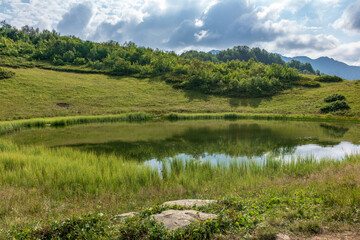 Fototapeta na wymiar Mountain lake on the green top of the slope with the sky reflected in it with clouds.