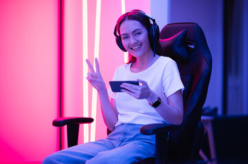 Young female professional cybersport gamer play mobile game by smartphone on eSport tournament online in colorful neon lights room. - obrazy, fototapety, plakaty