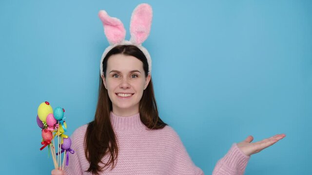 Pleased caucasian young woman in image of friendly Easter rabbit, raises palm as if showing something, gestures over blank space, holds small eggs, isolated on blue studio background. Advertisement