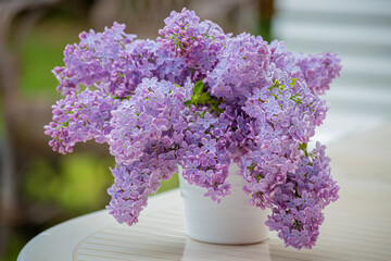 Lilacs syringa of bouquet in a white ceramic vase on a white table in rainy weather. Blurry background. Copy space. 