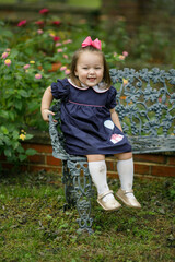 A toddler little girl with a navy dress and pink bow is sitting on an iron vintage bench