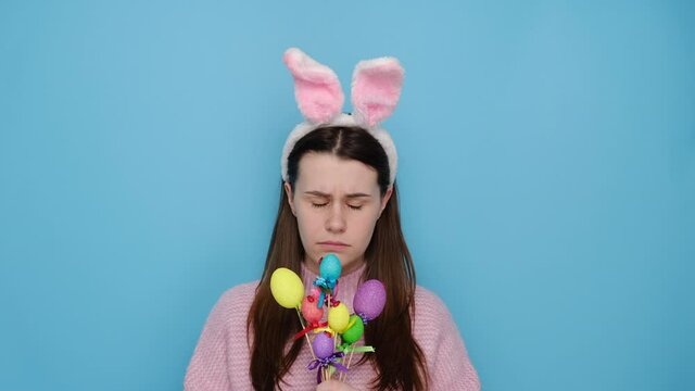 Discontent woman smirks face and looks unhappily, has spoiled day, poses with small Easter decorated colorful eggs, wears pink sweater, rabbit ears, poses with gloomy expression, isolated on blue wall