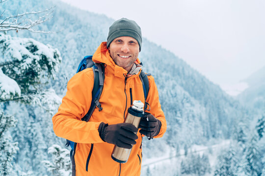 Laughing Man dressed bright orange softshell jacket with a hot drink thermos flask looking at camera while he trekking winter mountains route. Active people in the nature concept image.