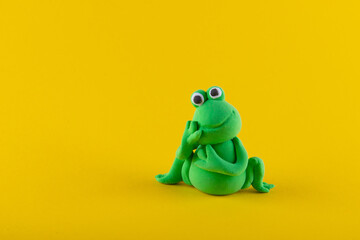 Frog toy figure made of light clay modeline sitting and holding head with hand on yellow background