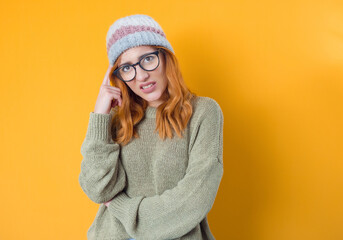 Pensive young woman, isolated on yellow background. Girl frowning her face holding finger on temple being in confusion with crossed arms