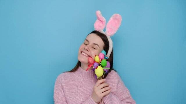 Good looking optimistic young woman with bunny fluffy ears, has a happy look, holds painted eggs on sticks, dressed in pink sweater, stands over blue studio background. Spring holiday concept