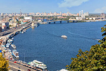 A landscape of summer Kyiv with a view of the Dnipro embankment in the old Podil district, a river station, piers, river trams and pleasure boats.