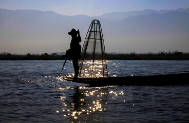 Silhouette of burmese traditional fisherman and basket standing on boat with paddle in the evening sun with glittering water reflections, blurred mountains background - Inle Lake, Myanmar