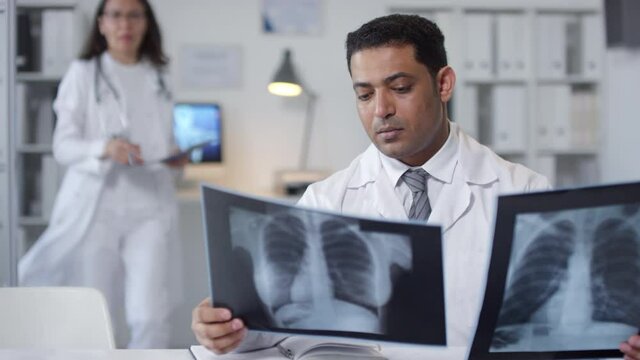 Portrait shot footage of serious Middle Eastern doctor sitting at desk in office room looking at X-ray pictures then discussing something with colleague
