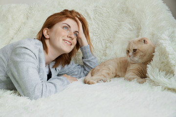 Happy dreamy young red-haired female lying on white comfortable plaid with ginger cat fluffy pet at home