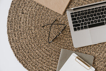 Home office desk workspace with laptop, clipboard, glasses on straw rattan background. Flat lay,...