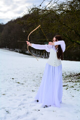 Young girl in long dress is standing in the snow with a bow and arrow