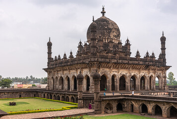 Fototapeta premium Vijayapura, Karnataka, India - November 8, 2013: NW corner view on brown stone Ibrahim Rauza mausoluem under gray cloudscape. Some green lawns.