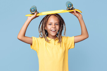 Positive little boy with african dreads holding skateboard on the head over blue background.