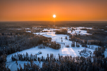 Warm sunrise over snowy countryside landscape. Pine forest covered in glowing snow. Drone aerial view.