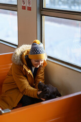 Fototapeta na wymiar Young happy caucasian woman wear winter clothes and hat sitting in local train, talking with her lovely dog, traveling together. Love pets. Vertical