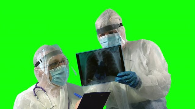 Two Physicians In PPE Suit Uniforms, Wearing Face Shields Are Inspecting A Patient's X-ray Of Lungs As A Test On Covid And Making Notes On Green Background