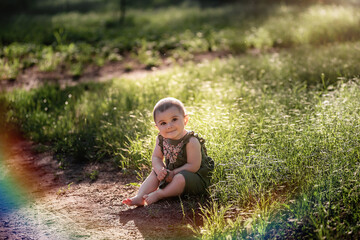 A little cute baby with short hair, in a green dress sits on the lawn in the grass near the path, looks up into the sky in the sun. She smiles at her family. Happy childhood, walk in the park. Summer