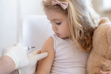 Immunization for children concept. Happy little cute blonde girl holding a toy and getting a flu shot not afraid of the syringe needle. Doctor injecting brave child with Covid-19 vaccine at clinic or