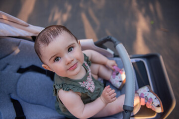 A cute little baby girl with green eyes in a dress sits in a stroller among a green park. Close-up...