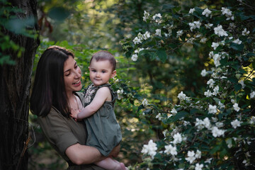 A young woman holds a little baby in her arms. A beautiful mother walks with her daughter in a green park near the jasmine bushes. Motherhood concept, love is limitless. Close up portrait, copy space