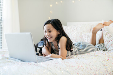 Smiling girl laying on bed with Boston Terrier home schooling on laptop computer