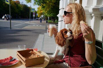 A 40-year-old blonde woman on the street at a table drinks coffee, next to her is a Cavalier King Charles Spaniel dog in a comic situation
