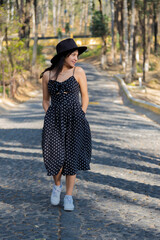 Hispanic woman with black dress walking in the middle of the street in the natural park - young woman for a walk in the middle of nature on a path of stones and trees enjoying the sunset sun