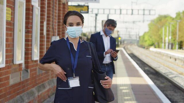 Portrait of nurse wearing uniform wearing face mask on standing on train station platform with other commuters- shot in slow motion