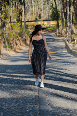 Hispanic woman in black dress walking in the middle of the street in the natural park - young woman for a walk in the middle of nature on a path of stones and trees