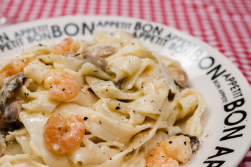 Delicious fettuccine with cream sauce of mushrooms and shrimp on a plate that says Bon Appetite with red checkered tablecloth.