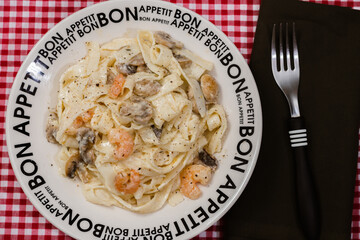 Delicious fettuccine with cream sauce of mushrooms and shrimp on a plate that says Bon Appetite with red checkered tablecloth.